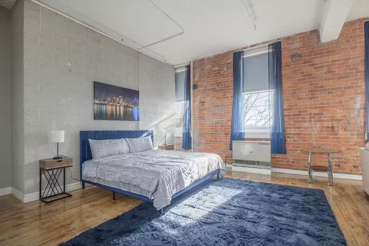 A bedroom with a bed, a nightstand, a lamp, a window with curtains, a brick wall, and a cityscape painting.
