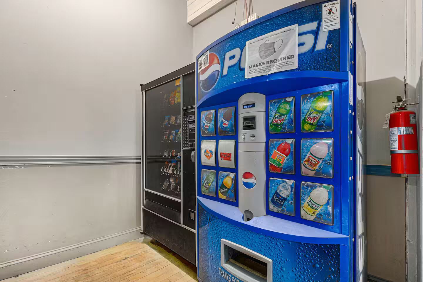 A vending machine with a variety of beverages and snacks, a fire extinguisher mounted on the wall, and a door with a frosted glass panel.