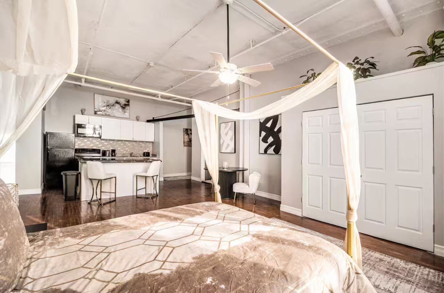 A bedroom with a canopy bed, a kitchenette in the background, and a white door.