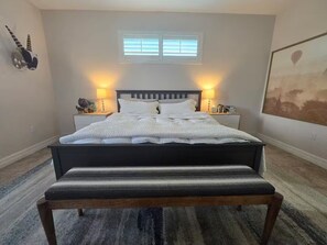 Un lit bien fait, recouvert d’une couette blanche, deux tables de chevet avec des lampes, une fenêtre avec des stores et un tableau encadré au mur.