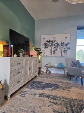Un salon avec une commode blanche, un téléviseur à écran plat, une grande fresque murale, un tapis à motifs et un fauteuil gris.