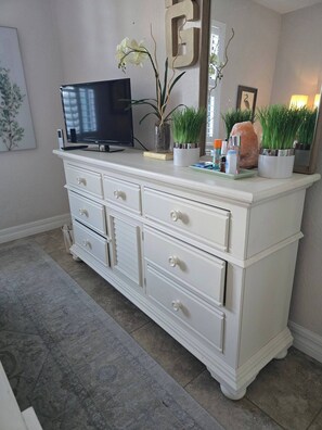Uma cômoda branca com uma TV de tela plana em cima, várias gavetas e plantas decorativas.