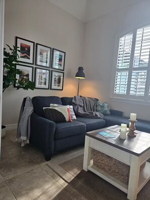 Ein Wohnzimmer mit einer Couch, einem Couchtisch, einer Lampe, gerahmten Bildern an der Wand und einem Fenster mit Jalousien.