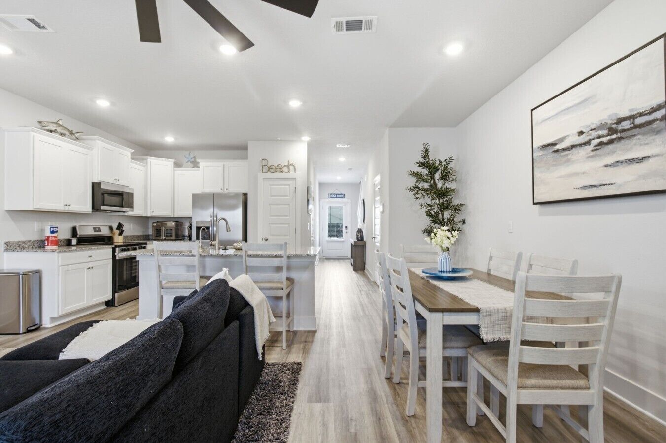A modern kitchen with white cabinetry, a dining area with a wooden table and white chairs, and a living room with a dark sofa.