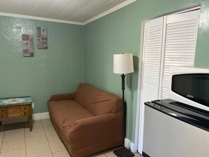 Una habitación con un sofá marrón, una mesita de madera, una lámpara de pie y un refrigerador blanco.