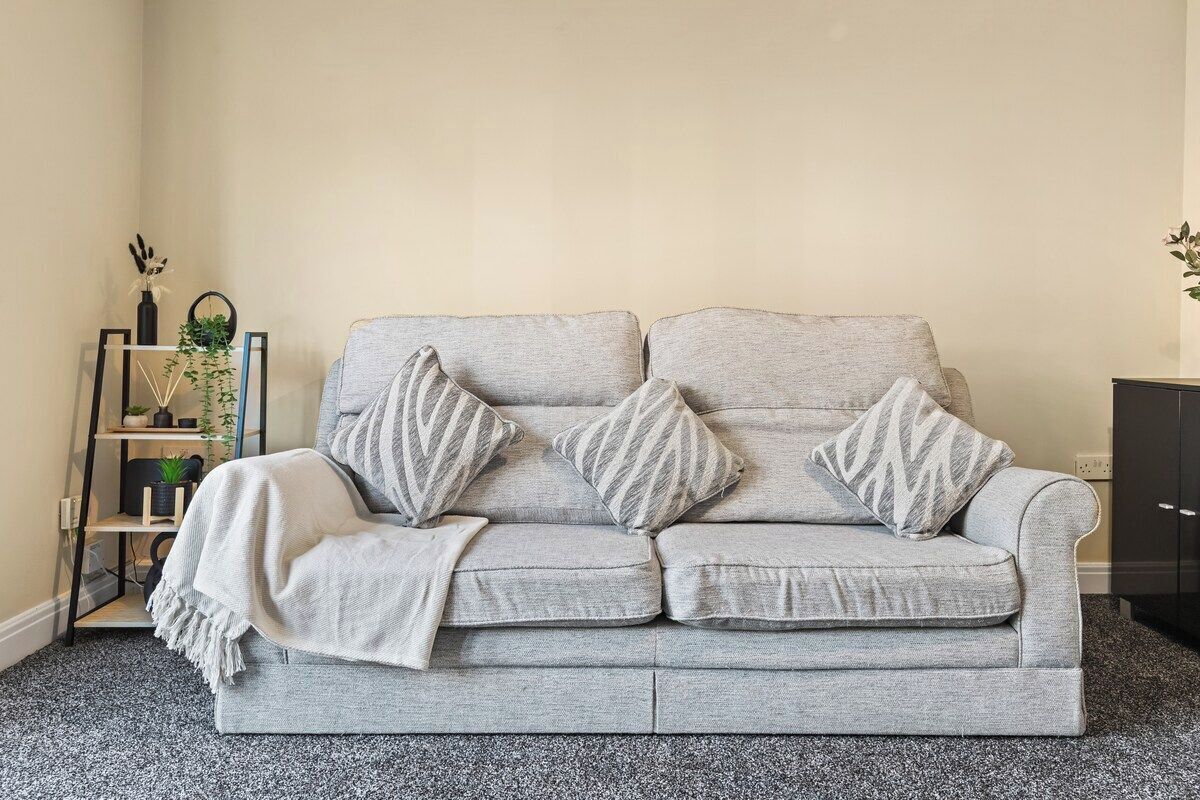 Um sofá cinzento com almofadas listradas e um cobertor, colocado em frente a uma parede com uma pequena prateleira e objetos decorativos.