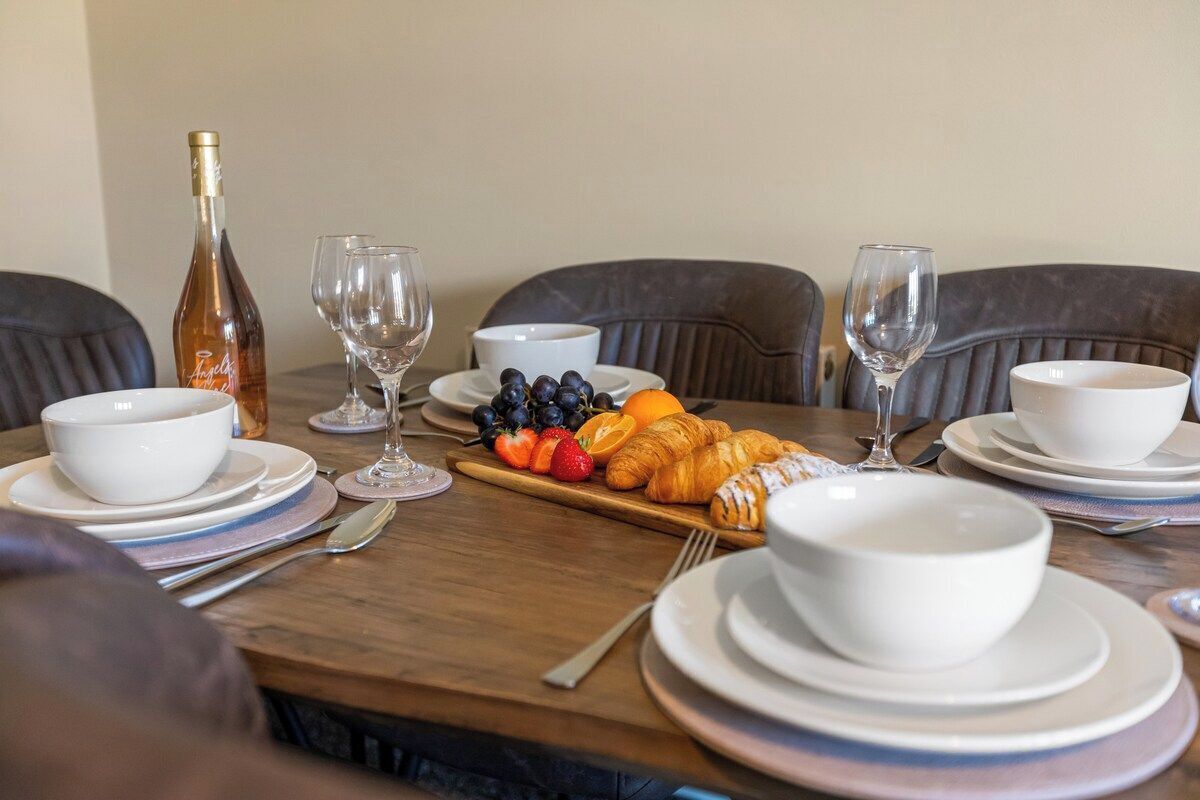 A dining table set with white bowls, plates, and cutlery, accompanied by a bottle, wine glasses, and a selection of fruits and pastries.