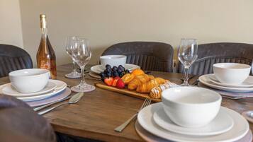 A dining table set with white bowls, plates, and cutlery, accompanied by a bottle, wine glasses, and a selection of fruits and pastries.
