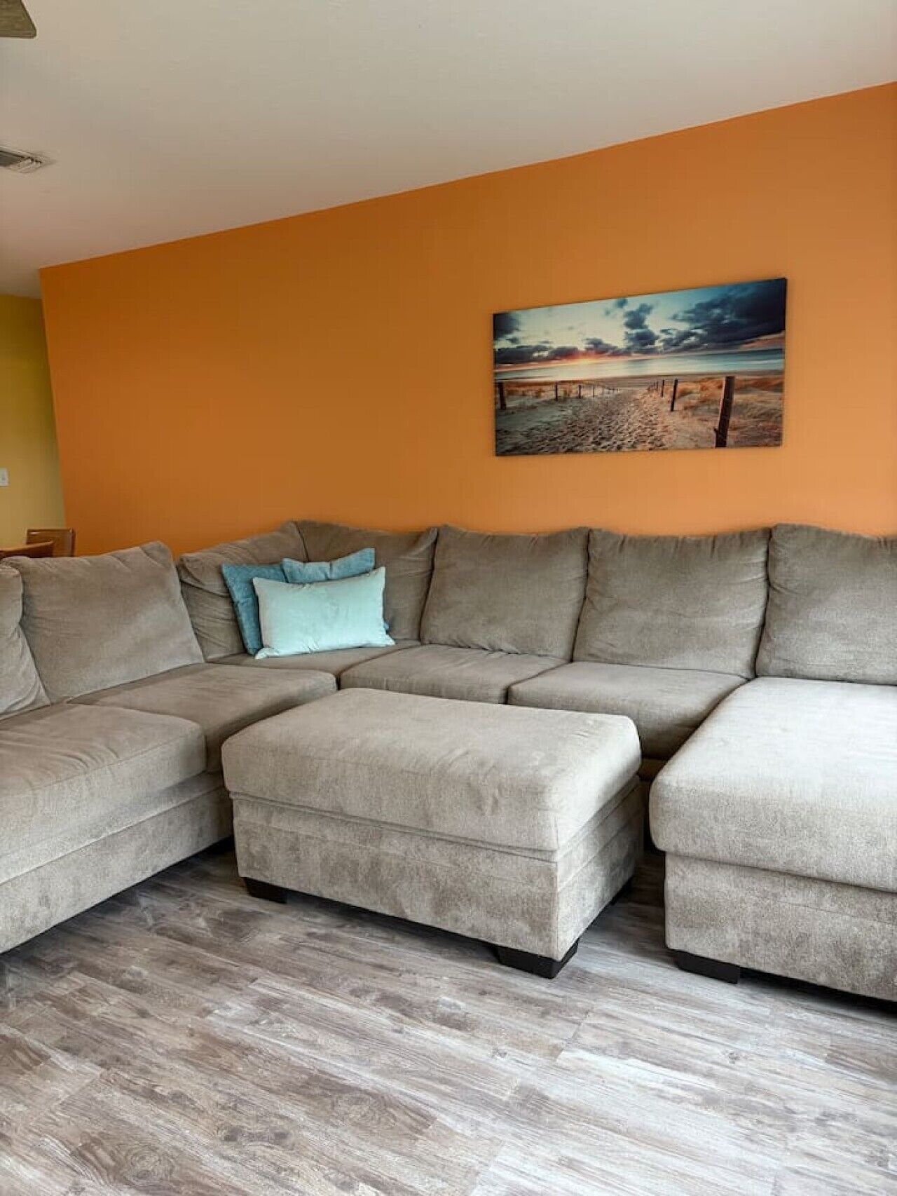 A living room with a sectional sofa, a small ottoman, and a painting on the wall.