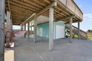 A covered outdoor area with a concrete floor, wooden beams, a blue building with a white garage door, and a staircase leading to a higher level. - Private Paradise w/Hot Tub @ The SeaBird (Galveston)
