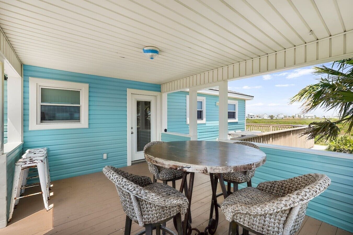 A covered outdoor area with a round table and wicker chairs, a door, and a view of the beach.