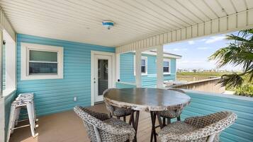 A covered outdoor area with a round table and wicker chairs, a door, and a view of the beach.