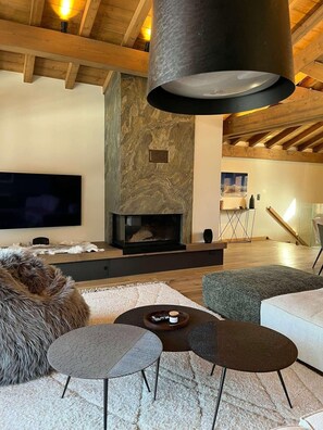 A modern living room with a stone fireplace, wooden beams, a flat-screen TV, and a cozy seating area with a fur pouf and two round tables. - 3rd floor, terrace, fireplace or stove, tv, 157m², Peisey-Vallandry (Peisey-Nancroix)