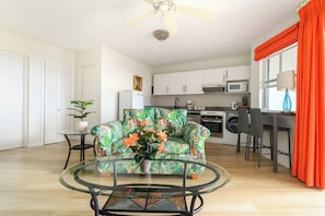 A modern living room with a glass coffee table, a sofa with a tropical print, and a kitchen area with white cabinets and a microwave. - Rum Fun Sun at Lantana No. 25 | Refreshed 1BR, 2 Pools, West Coast Barbados (St. James)