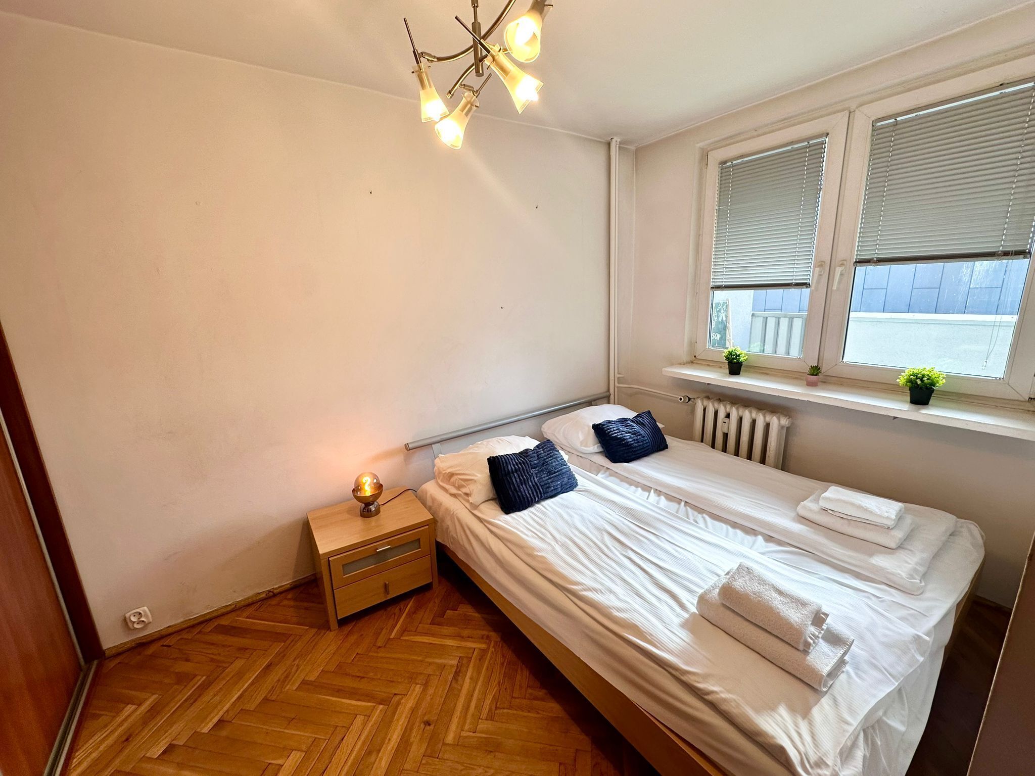 Een slaapkamer met een tweepersoonsbed, nachtkastjes, een radiator, een raam met zonwering en een kleine plant op de vensterbank.