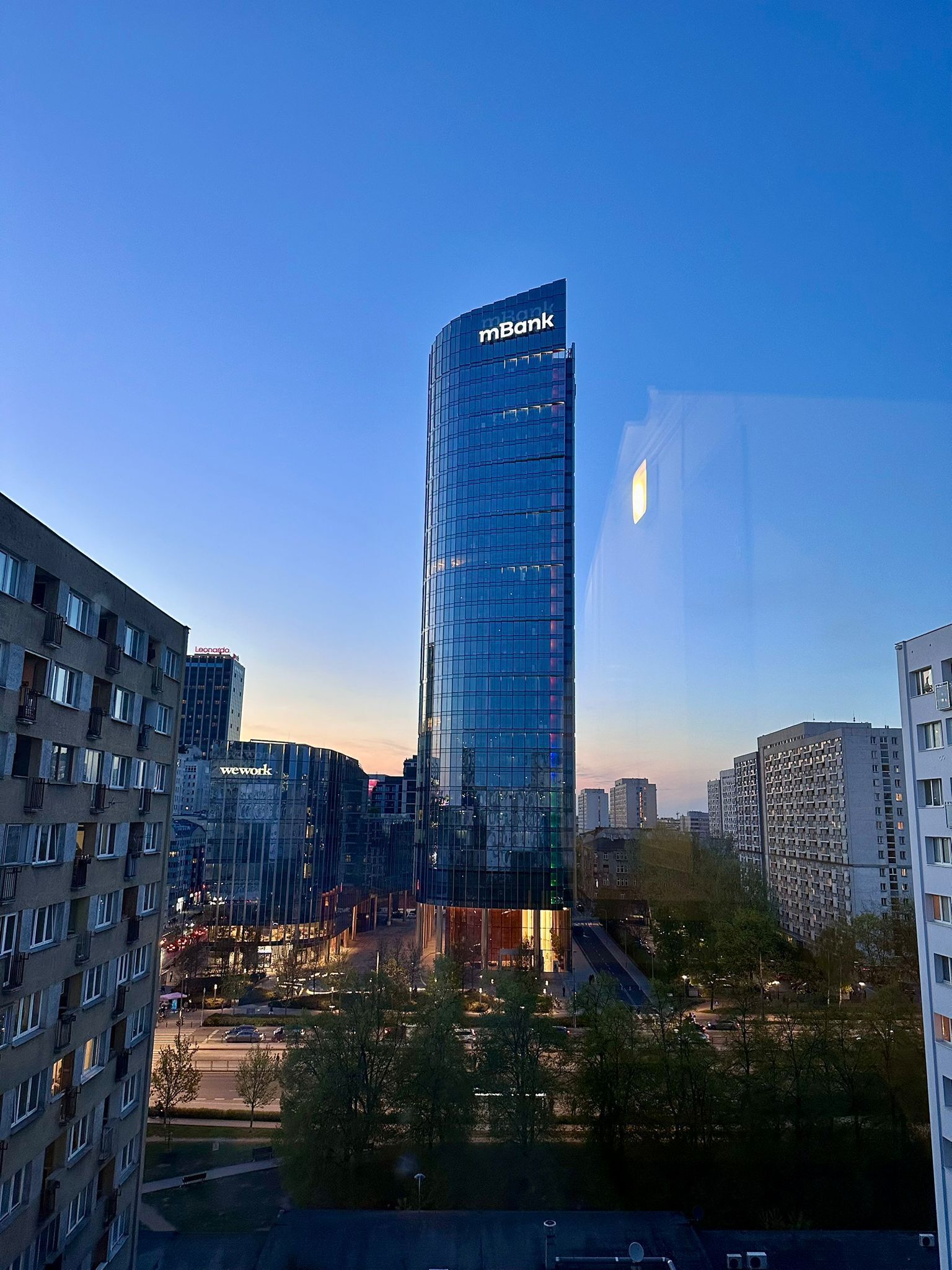 A modern high-rise building with a glass facade, surrounded by other buildings and a park area.