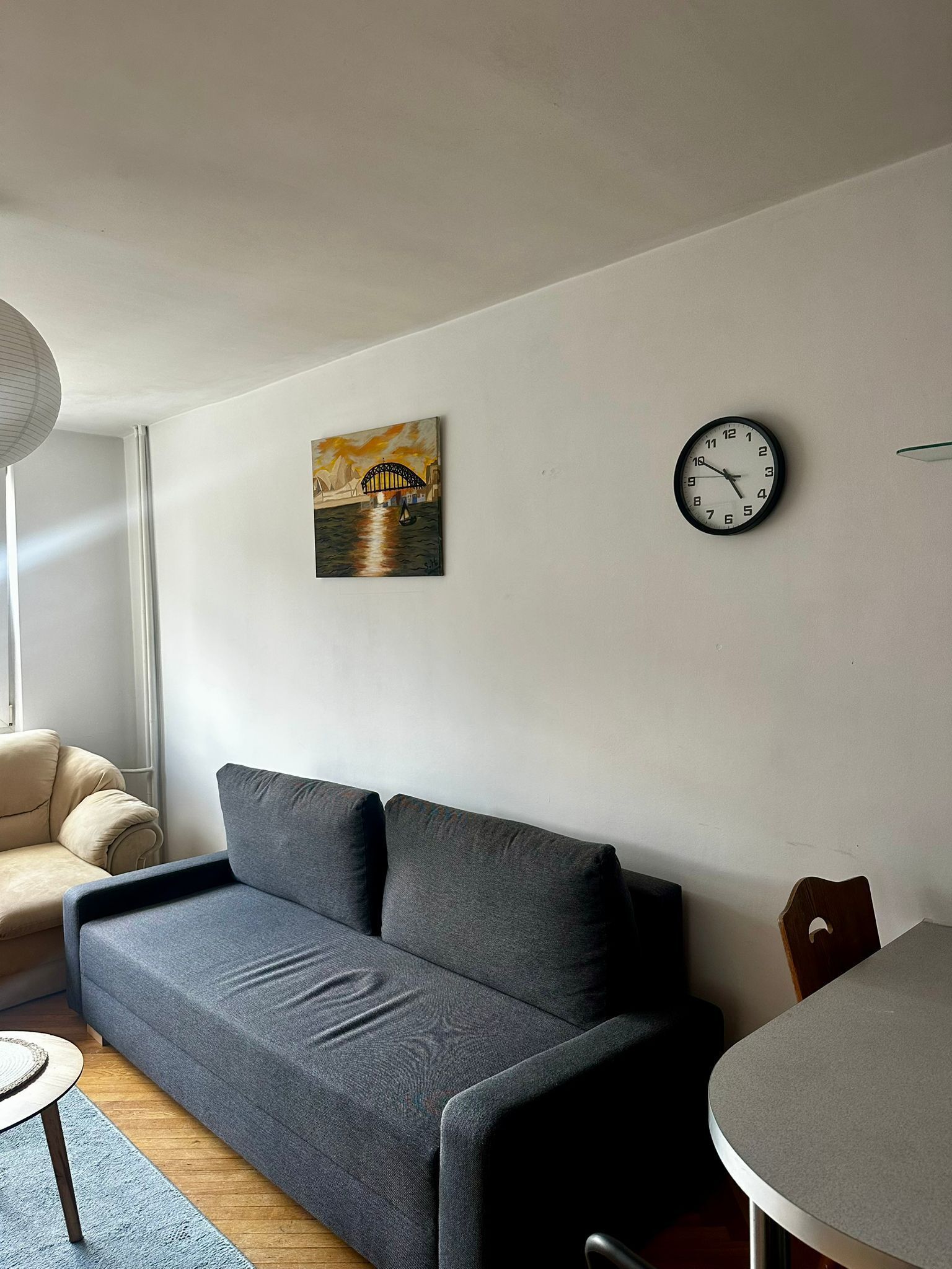 A modern living room with a grey sofa, a painting of a bridge, and a wall clock.