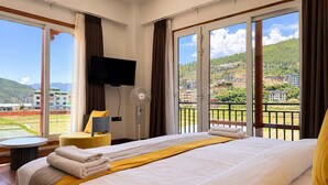 A hotel room with a large bed, a flat-screen TV, a fan, and a view of a mountainous landscape. - Golden Paddy Field  (Paro)
