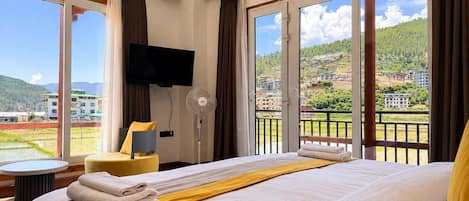 A hotel room with a large bed, a flat-screen TV, a fan, and a view of a mountainous landscape.
