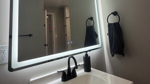Une salle de bain avec un lavabo blanc, un grand miroir doté d’un éclairage intégré et un porte-serviettes fixé au mur.