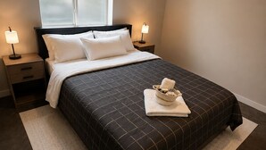 Un lit bien fait, recouvert d’une couverture sombre, deux lampes de chevet, une fenêtre à vitres dépolies et une petite table de chevet avec une lampe.