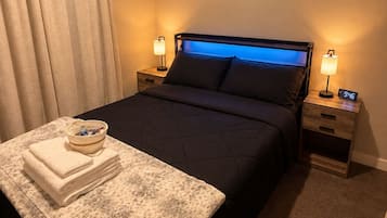 A hotel room with a bed, two bedside tables, a nightstand, a lamp, a clock, a window with a view, and a towel stack.
