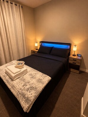 Une chambre d’hôtel avec un lit, deux tables de chevet, une commode, une lampe, une horloge, une fenêtre donnant sur un beau paysage et une pile de serviettes.