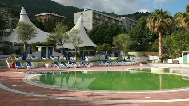 Una piscina con lettini e ombrelloni, circondata da edifici e palme.