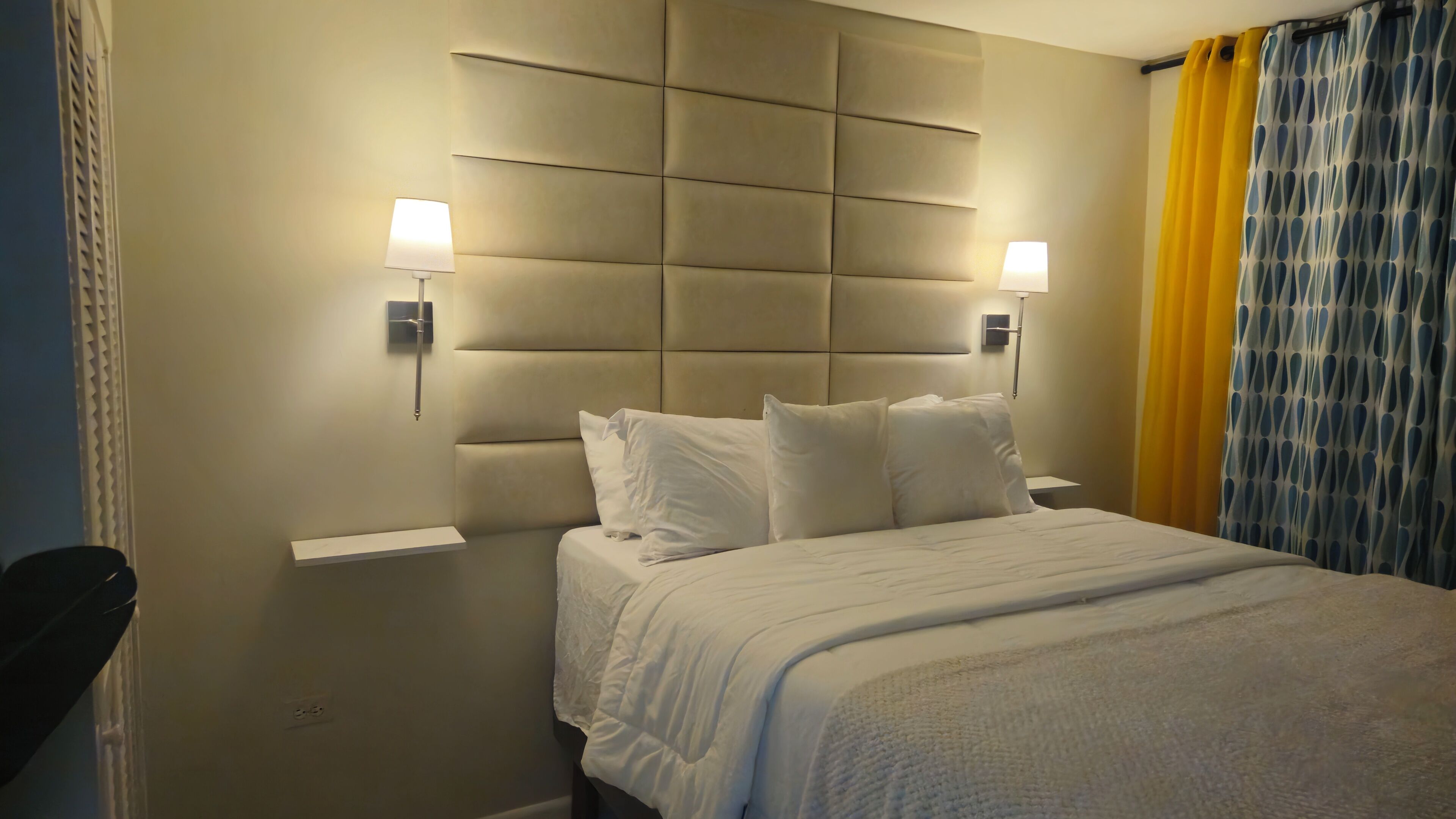 Ein Hotelzimmer mit einem großen Bett, zwei Nachttischlampen, einem gepolsterten Kopfteil und einem Vorhang mit geometrischem Muster.