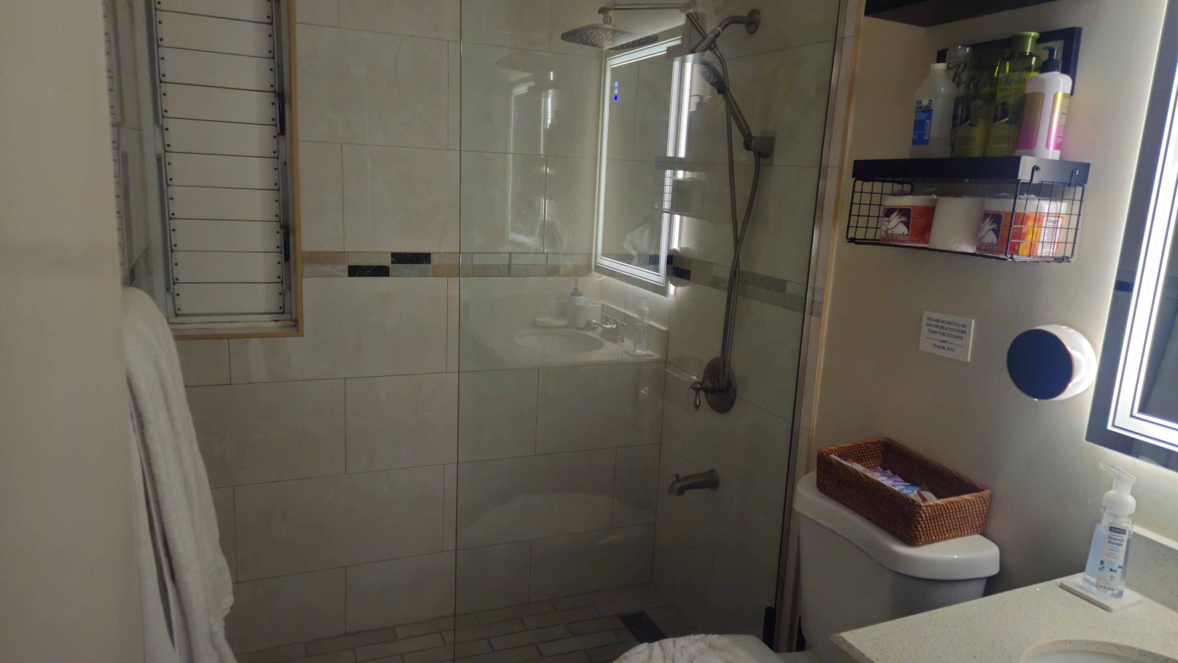 A bathroom with a glass-enclosed shower, a toilet, and a sink. There are toiletries on the wall and a basket on the toilet.