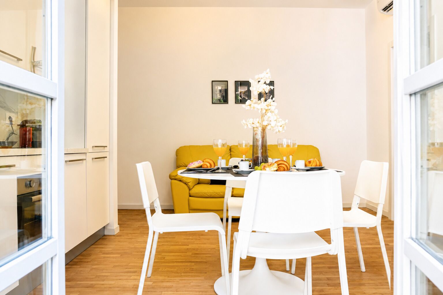 Een eethoek met een tafel voor vier personen, stoelen, een gele bank en een vaas met bloemen.