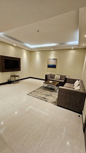 A spacious living room with a flat-screen TV mounted on the wall, a sofa set, a coffee table, and a patterned rug. - Best Level (Al Khobar)