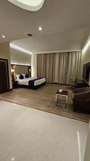 Habitación de hotel con cama, televisor, sofá, mesa de centro y una ventana grande con cortinas.