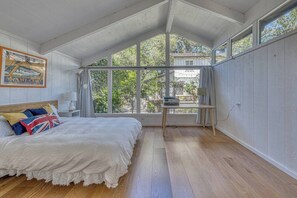 Ein Schlafzimmer mit einem großen Bett, einem Schreibtisch mit Lampe und Blick auf Grünflächen.
