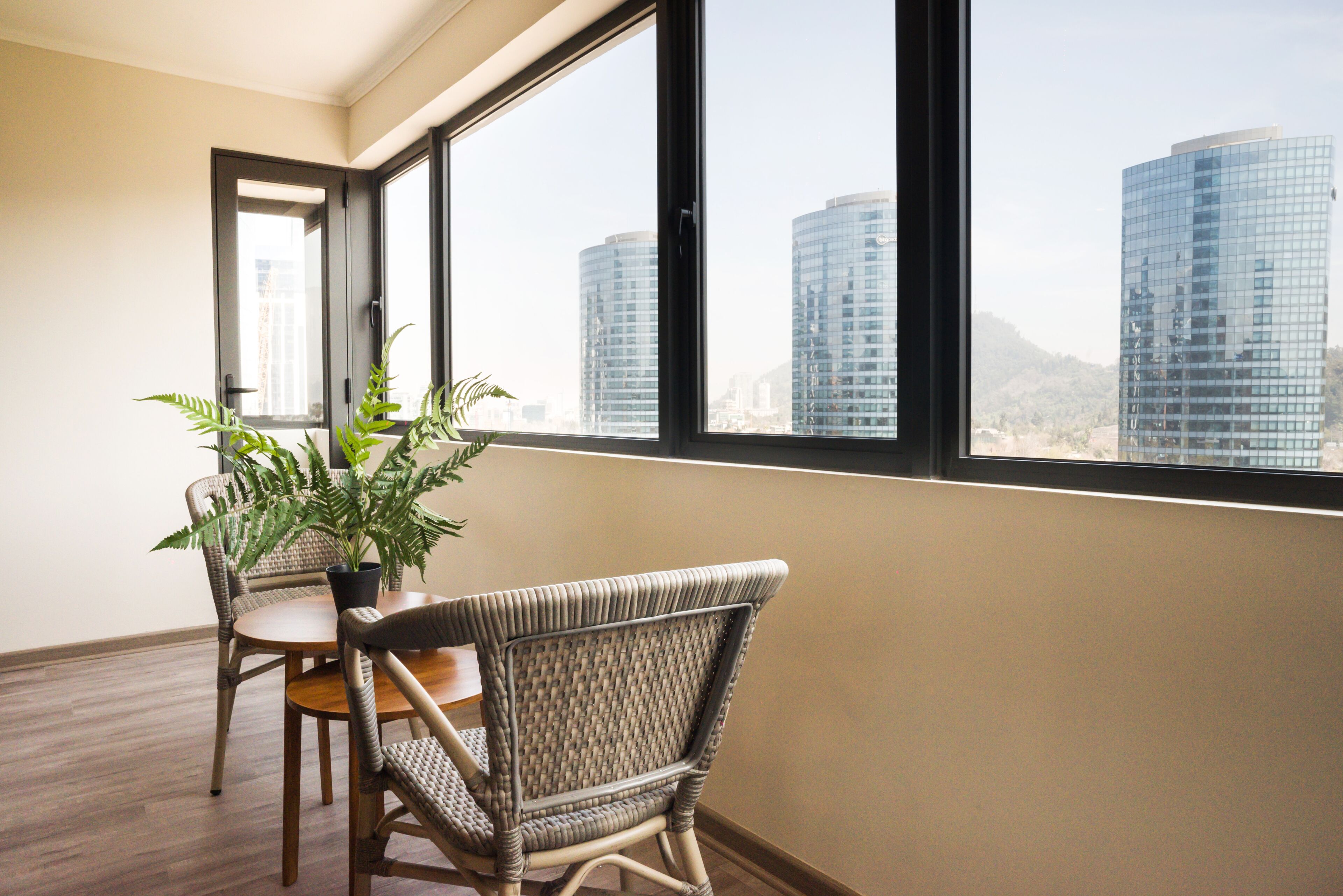 A room with a view of tall buildings through large windows, a wicker chair, a small table, and a potted plant.