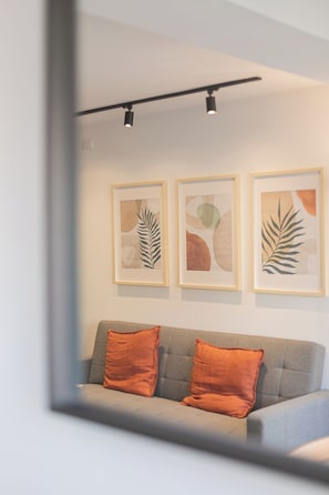 A modern living room with a grey sofa and two orange cushions, a wall-mounted light fixture, and three framed abstract art pieces. - Vitacura Apartments by Andes STR (Las Condes)