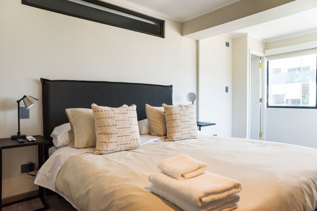 A hotel room with a bed, pillows, a bedside table with a lamp, and a window.