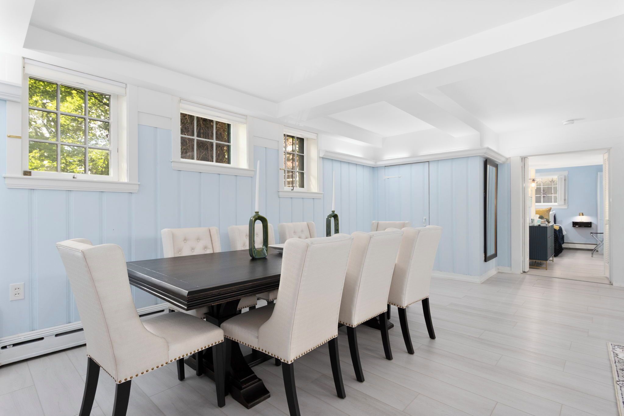 A dining area with a rectangular table and six chairs, a window with a view of trees, and a door leading to another room.
