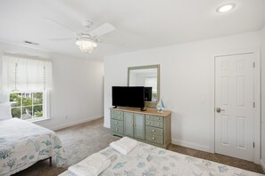 Une chambre à coucher comprenant un lit, une commode, un téléviseur, un ventilateur de plafond, une fenêtre avec des rideaux et une porte.