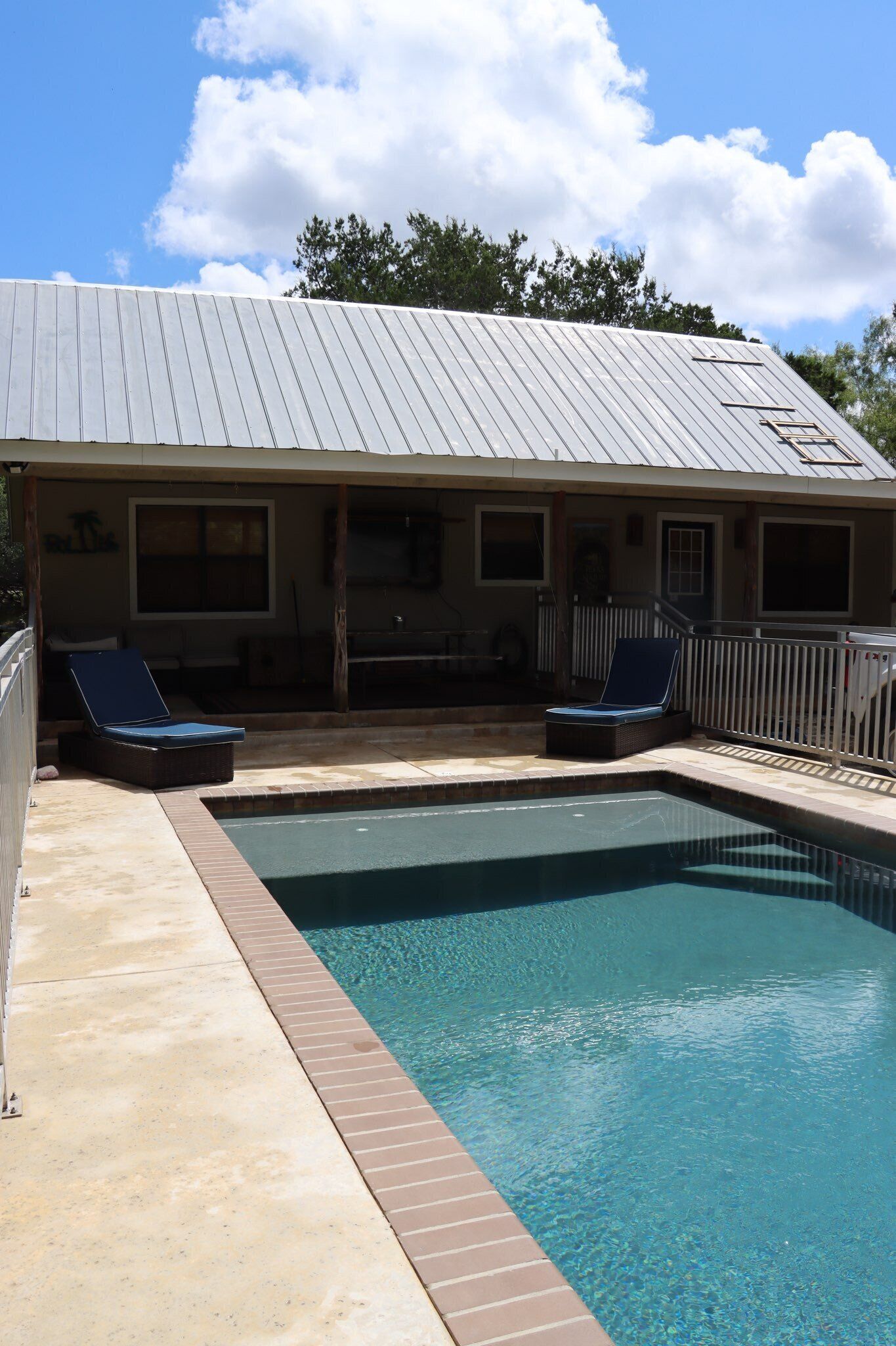 A swimming pool with two lounge chairs, a covered patio area, and a building with a metal roof.