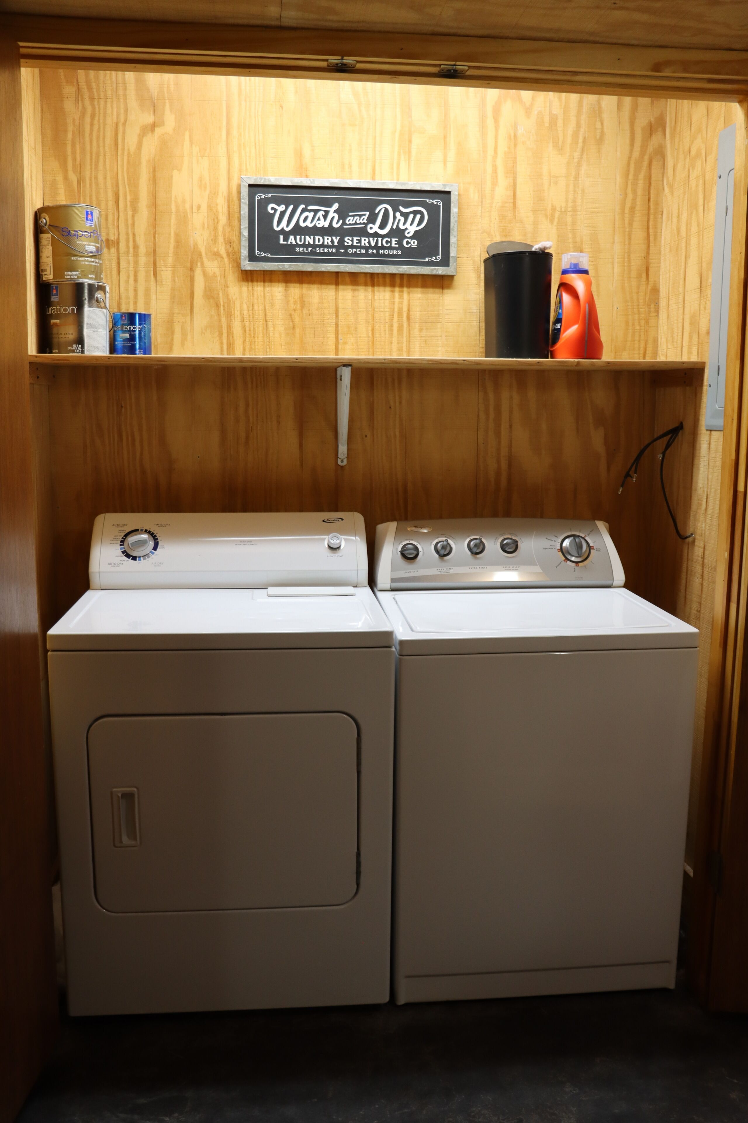 A laundry area with two front-loading washing machines, a shelf with detergent bottles, and a sign indicating a self-serve laundry service.
