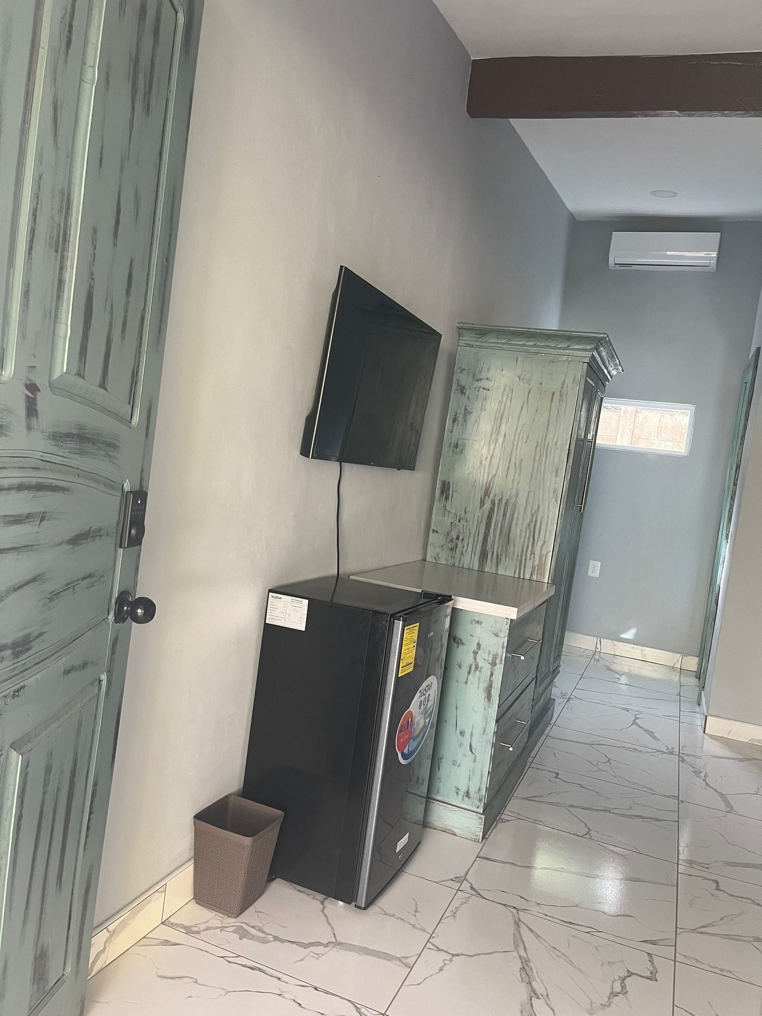 A hallway with a door, a trash bin, a black refrigerator, and a wall-mounted television.