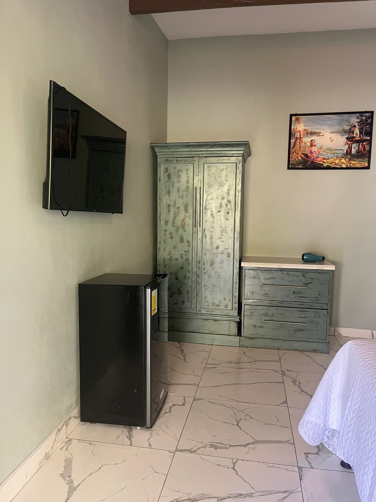 A room with a television mounted on the wall, a cabinet with drawers, a tiled floor, and a bed with a white cover.