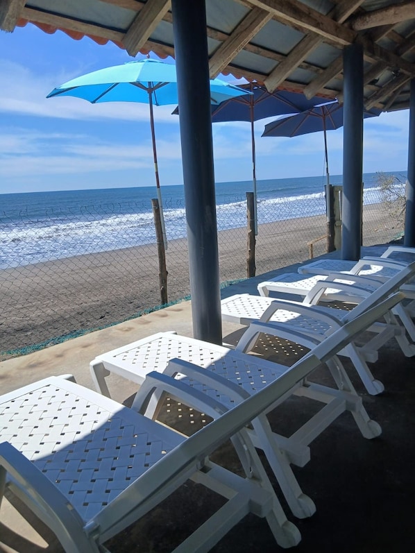 A beachside seating area with white plastic chairs and blue umbrellas, overlooking the ocean. - Boni Beach (Sonsonate)
