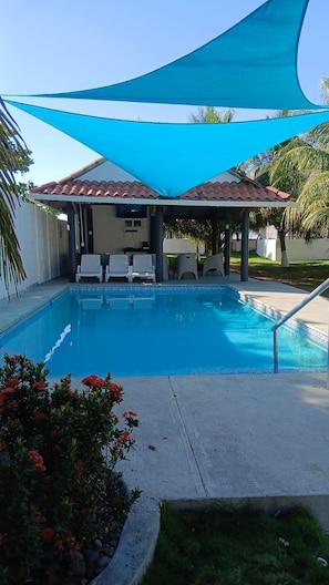 A swimming pool with a covered patio area, surrounded by greenery and a clear sky.