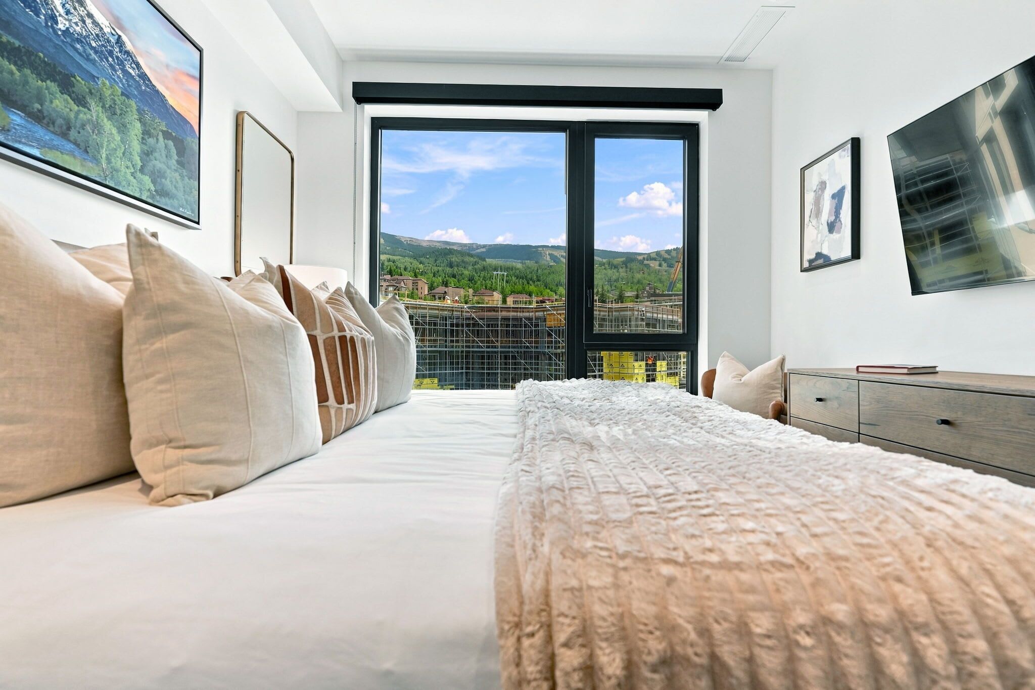 Un dormitorio moderno con una cama grande, vista a una obra en construcción y un televisor montado en la pared.