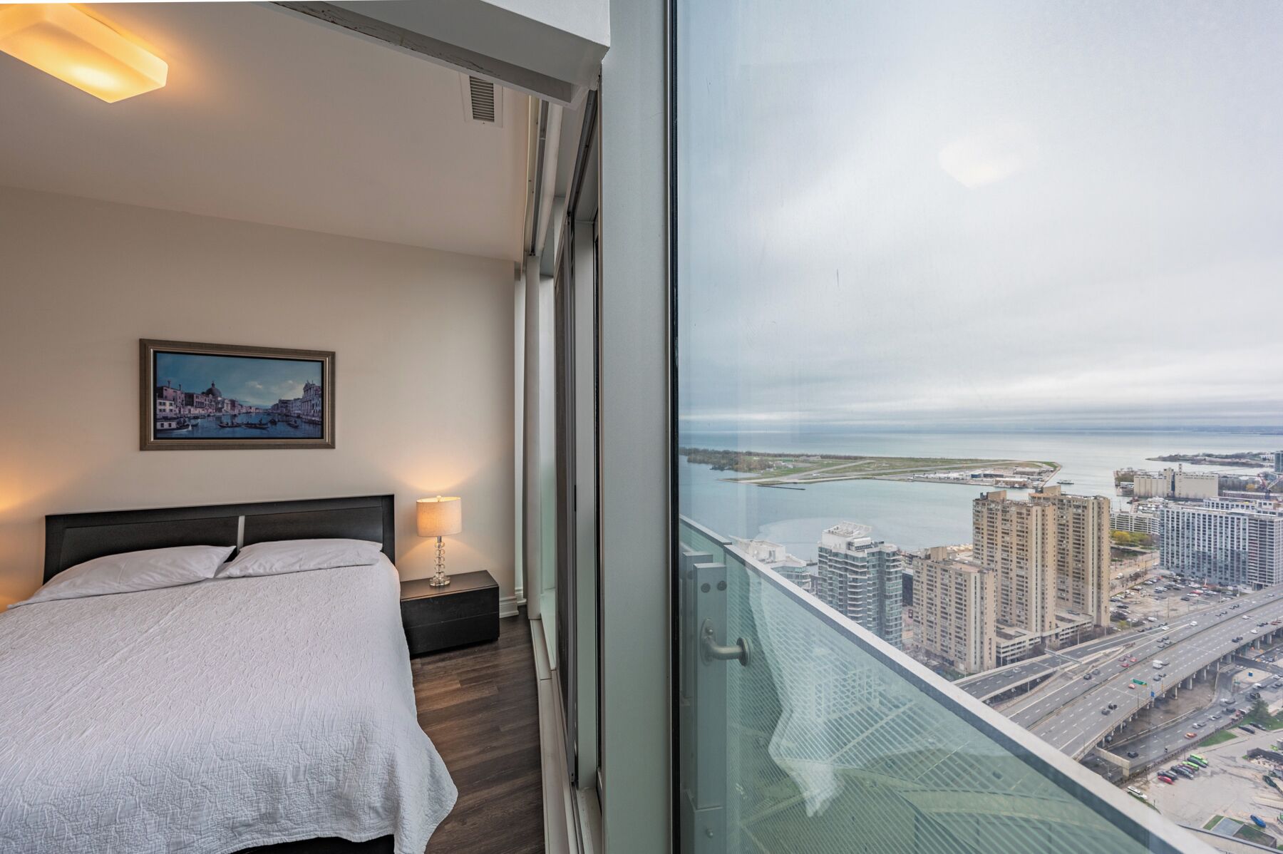 A bedroom with a bed, a nightstand, a lamp, and a city view through a large window.