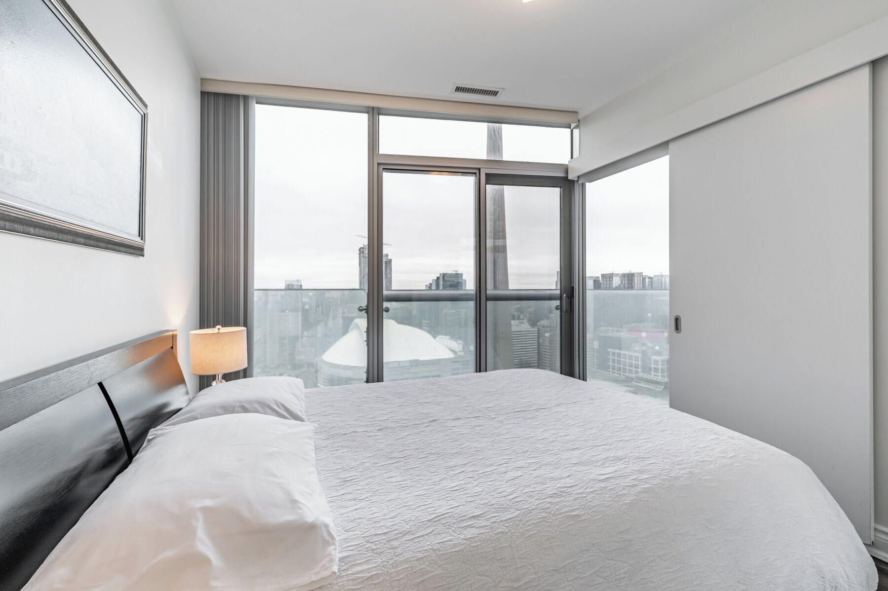 A hotel room with a large bed, a nightstand, a lamp, and a view of the city through floor-to-ceiling windows.