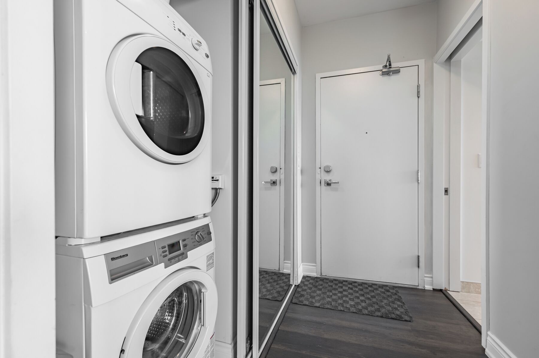 A laundry room with stacked washers and dryers, a door, and a doormat.