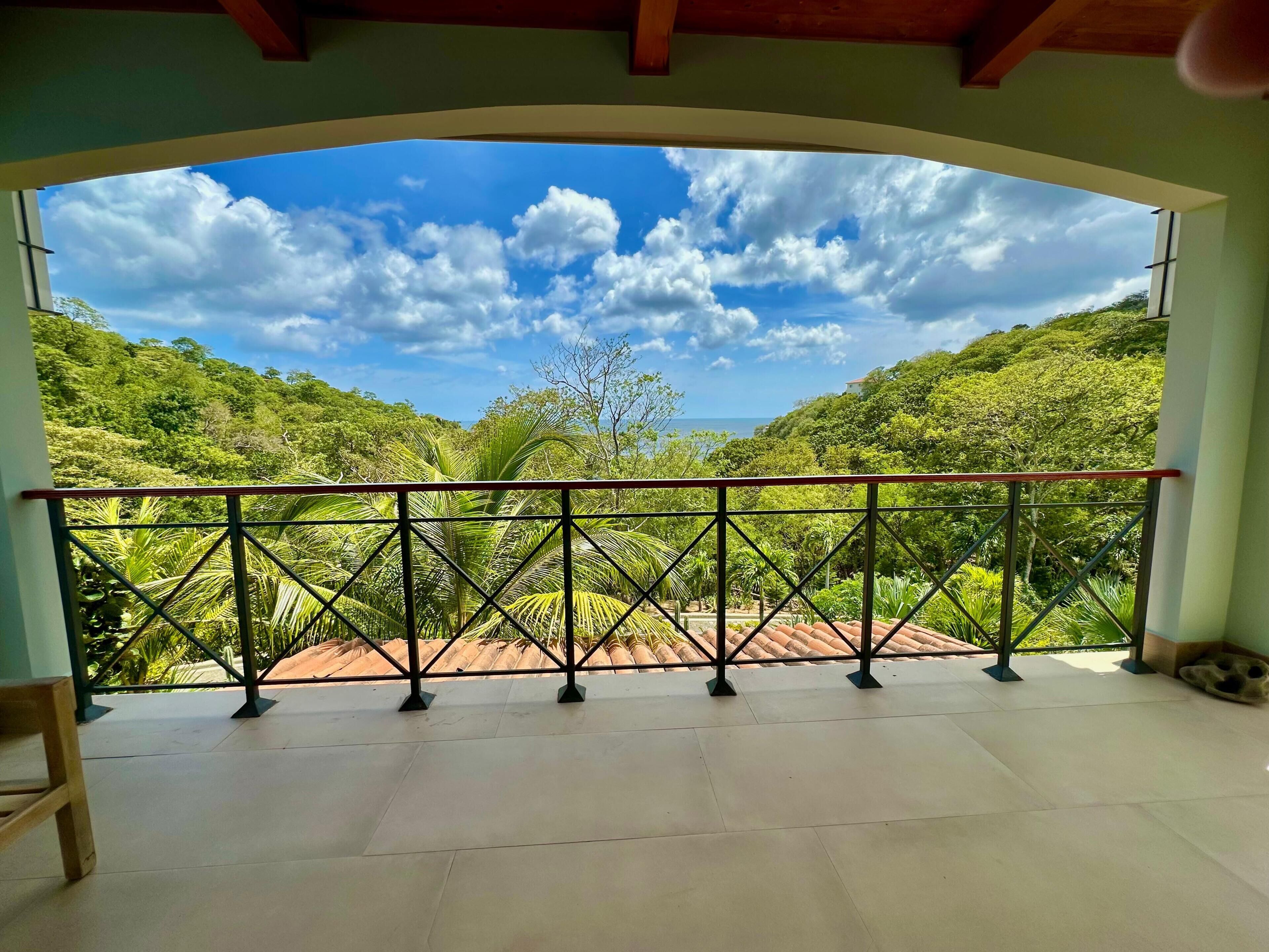 Un balcón con barandilla de metal, suelo de baldosas y vistas a un paisaje verde exuberante con árboles y cielo despejado.
